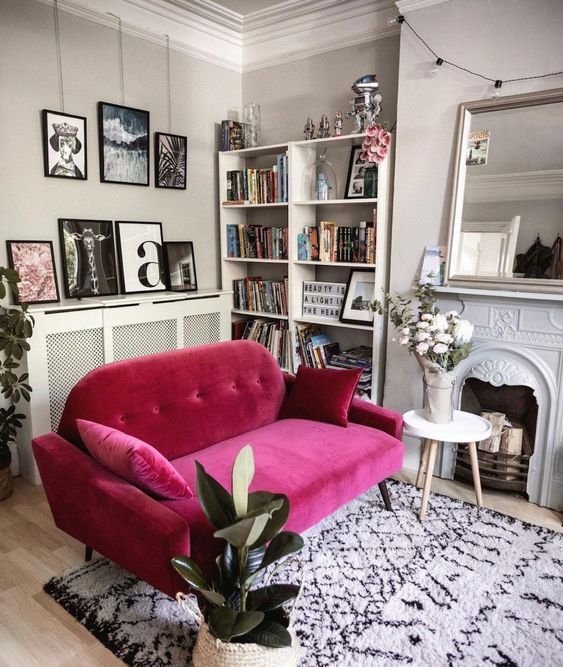 a neutral living room with a non working fireplace, a large mirror, a bookcase, a magenta sofa and some potted plants