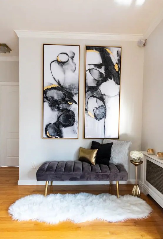 a glam and chic nook with a grey upholstered bench, statement watercolor artworks with gold, a gold pillow, legs and a small table