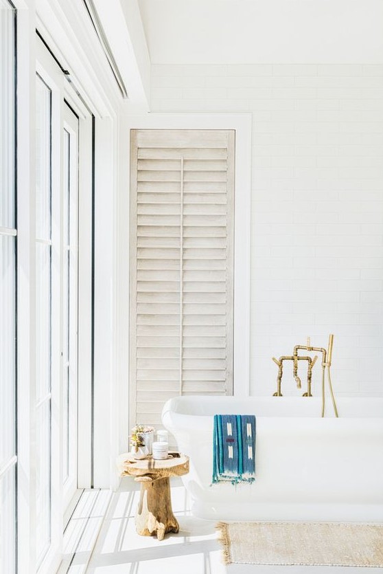a light-filled white bathroom with a free-standing tub, brass fixtures, a wooden stool and a glazed wall plus shutters
