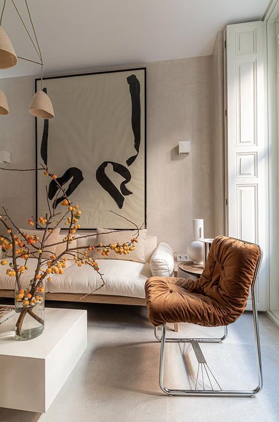 an adorable minimalist living room done in neutrals, with a terracotta upholstered chair, branches in a vase and tan chandelier