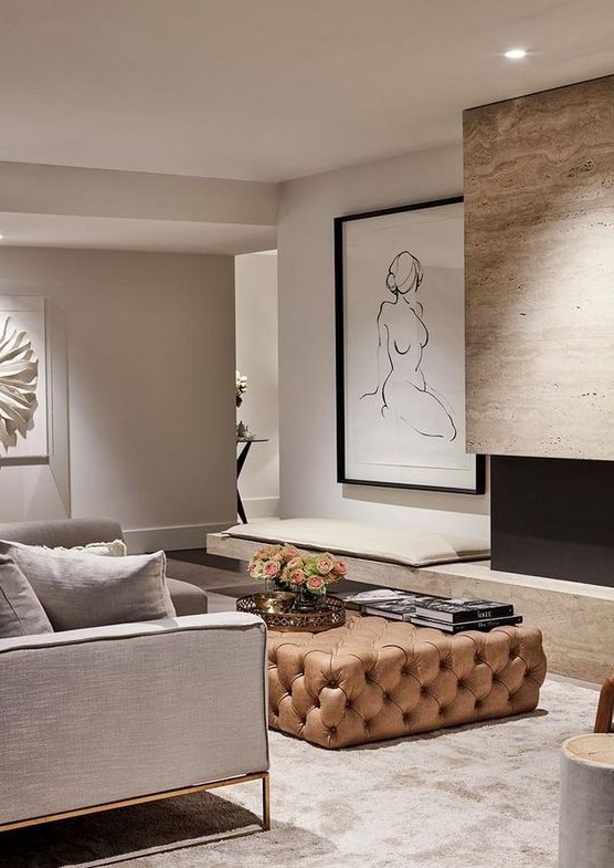 a gorgeous minimalist living room done in greys, with a tan fireplace and a light brown tufted ottoman for a sutble touch of color