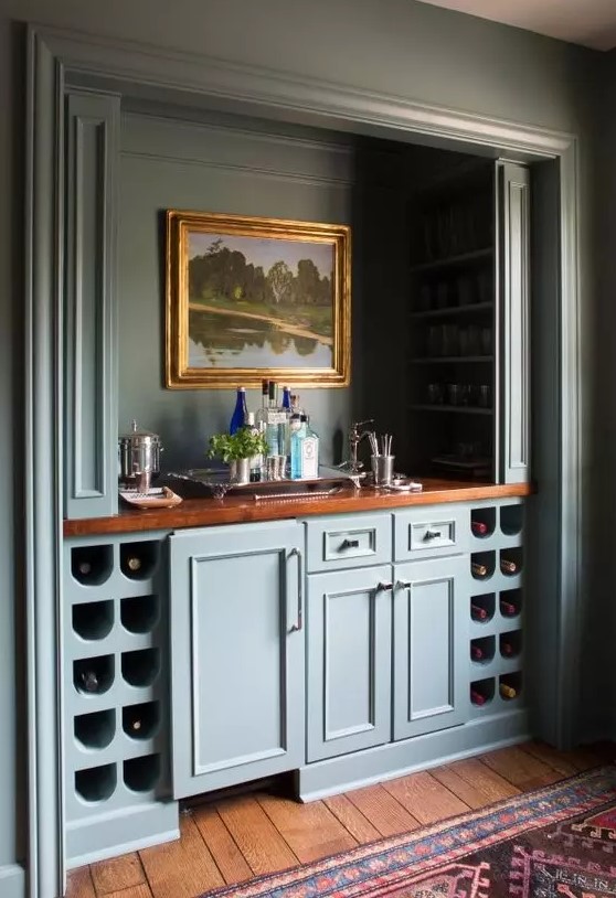 a vintage built-in home bar with blue cabinetry, wine storage compartments, artwork, wine bottles and wine glasses