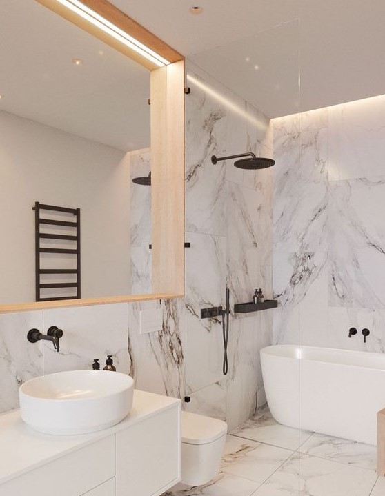 a refined white bathroom done with large scale marble tiles, with black fixtures and a large mirror in a wooden frame