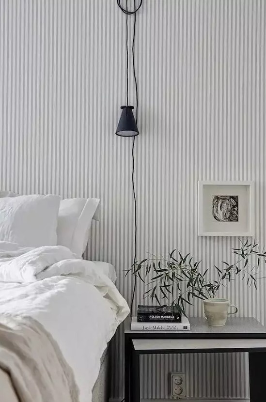 a Scandinavian bedroom finished off with a grey and white striped wall, a bed and a nightstand, some greenery and art is welcoming