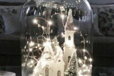 a Christmas cloche terrarium with a snowy scene with a church, a man and a dog and a Christmas tree plus lights