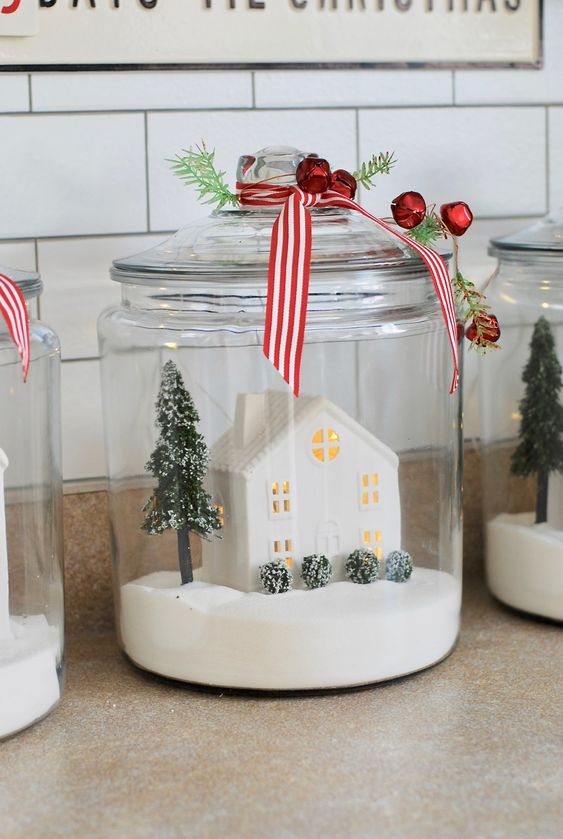 a Christmas terrarium with faux snow, a mini house, a Christmas tree and bushes topped with mini red bells