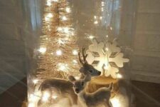 a cloche Christmas terrarium with faux snow, a couple of deer, a snowflake and a bottle brush tree plus lights