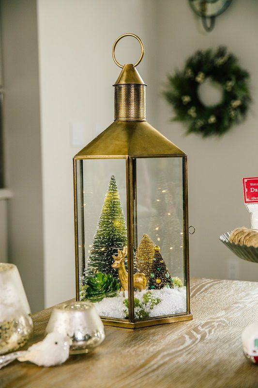 a lantern Christmas terrarium with faux snow, a gold deer, bottle brush trees and some greenery is a chic solution