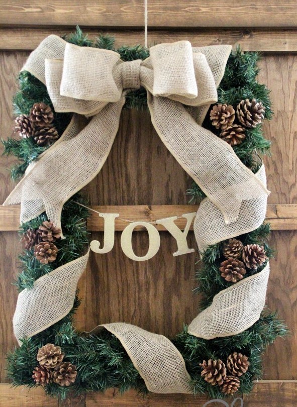 a rustic Christmas frame wreath with evergreens, pinecones, a burlap bow and ribbons interweaving it plus a letter banner