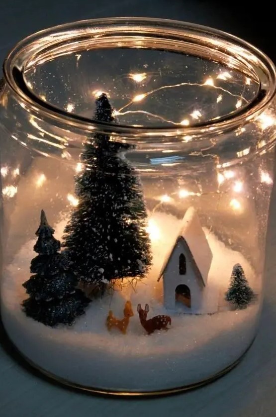 an oversized jar with faux snow, little deer figurines, a house and some bottle brush trees and lights is pure magic