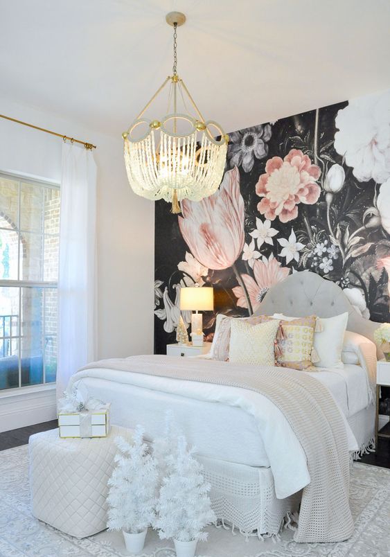 a beautiful bedroom with a dark floral accent wall, a grey upholstered bed with neutral bedding, an upholstered pouf and a beaded chandelier