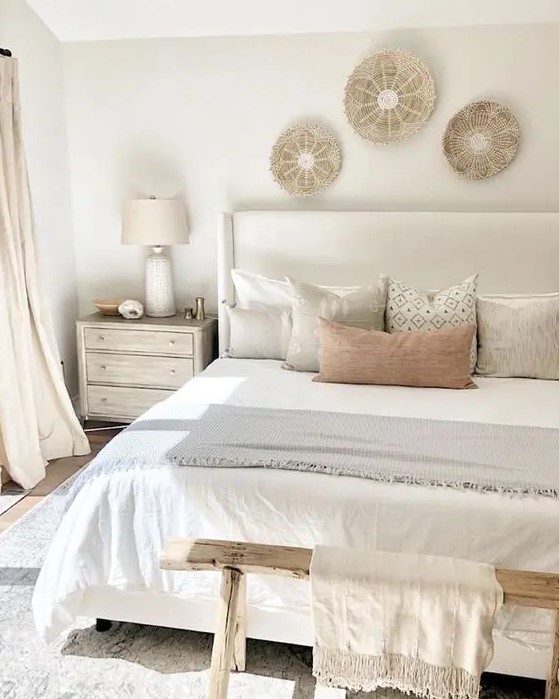 a neutral and warm bedroom with a creamy upholstered bed, a wooden bench, woven plates, printed pillows and neutral curtains