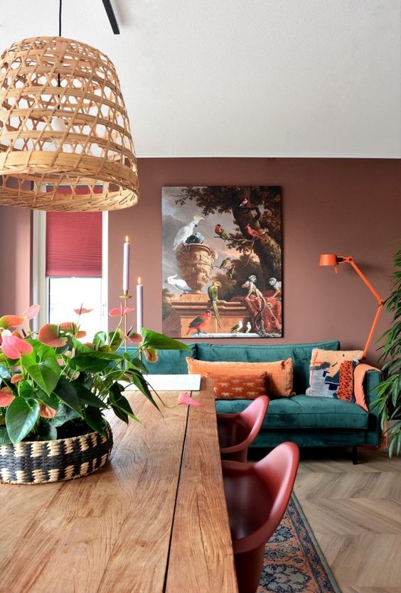 a beautiful muted-colored living room with chocolate brown walls, a green sofa with orange pillows and a lamp, an artwork and a dining zone