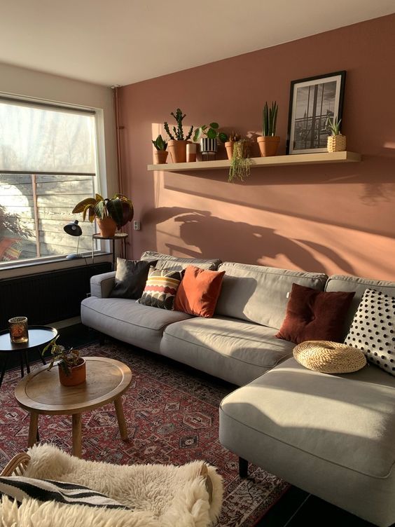 a pretty living room with a rust accent wall, a grey sofa with bright pillows, an open shelf, a bold printed rug and mini side tables