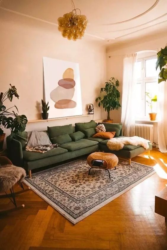 a muted and warm-colored living room with tan walls and a ceiling, a green sectional, a woven pouf, a printed rug and potted plants