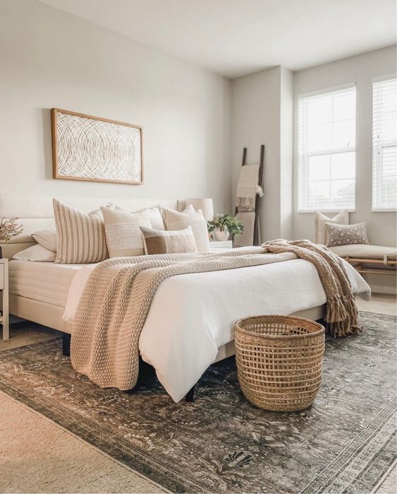 a cozy neutral bedroom with a bed and layered bedding, a basket, a printed rug, a bench with pillows and nightstands
