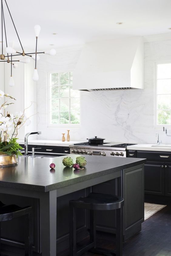a cool black kitchen with a large white hood, a large black kitchen island and matching stools, a chandelier and some brass touches