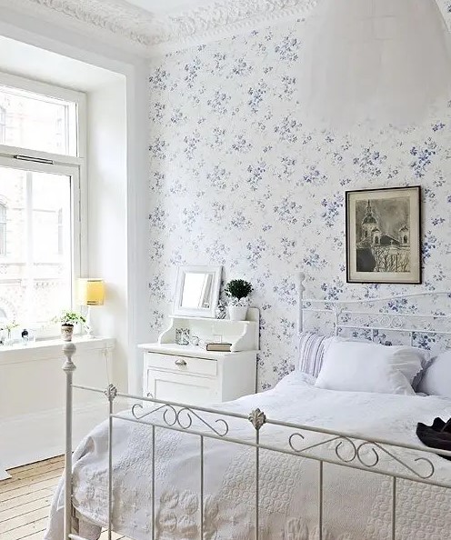 a vintage-inspired bedroom with a blue floral wall, a white forged bed, white furniture, a dark artwork and a pendant lamp