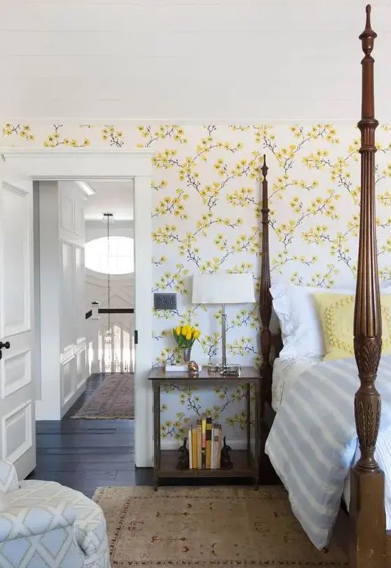 a vintage inspired bedroom with yellow floral walls, a heavy bed with pillars, a chair and nightstands plus blooms