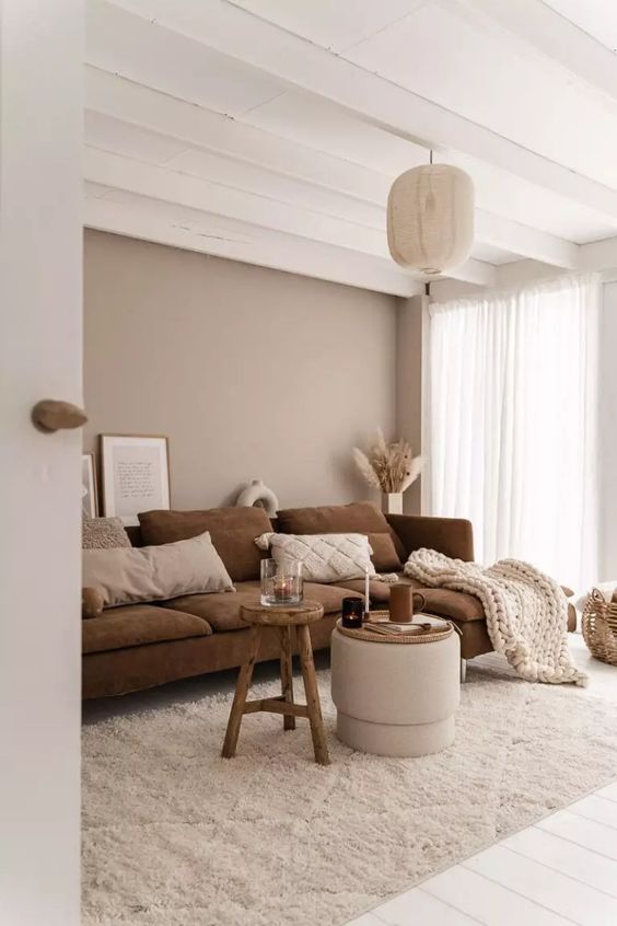 a warm-colored living room with a greige accent wall, a brown sofa with neutral pillows and a blanket, a stool and a pouf