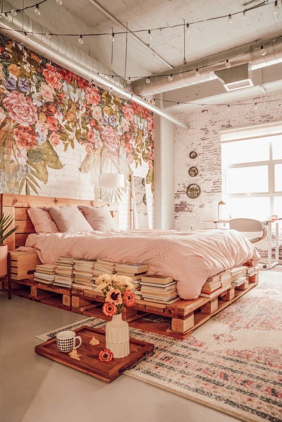 an eclectic bedroom with a painted floral wall, a pallet bed with blush bedding, stacks of books and bulbs over the space
