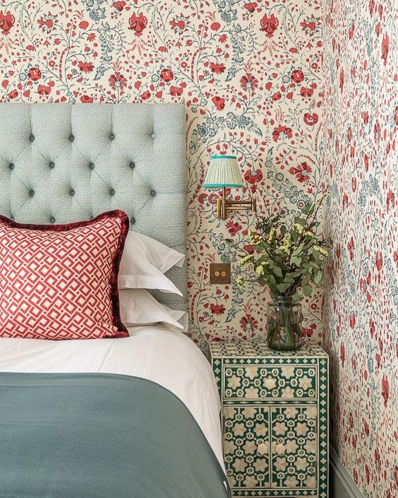 an eye catchy bedroom with bold blue and red floral wallpaper, a mint upholstered bed with red printed and green bedding, a green and white floral nightstand