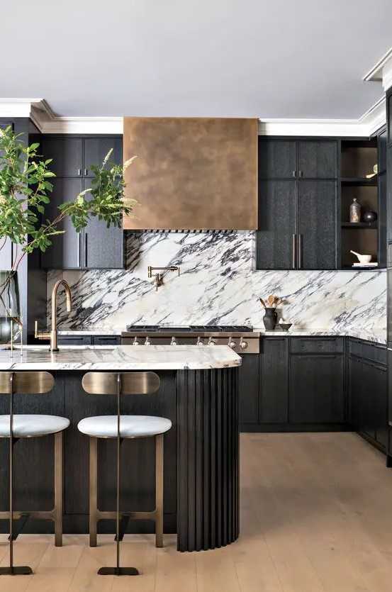 a sophisticated black kitchen with a white marble backsplash, an aged brass hood, a fluted curved kitchen island and tall stools with brass backs