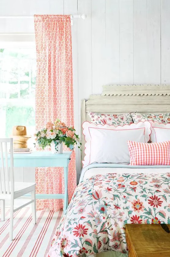 a bright summer bedroom with a vintage bed, vintage-inspired red, green and cream bedding with ruffled pillows, a blue desk and a white chair, printed curtains and a rug