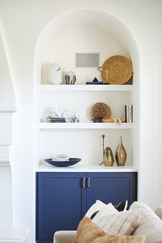 an arched niche with shelves that shows off stacks of books, baskets, vases and dishes plus a navy built-in cabinet for storage
