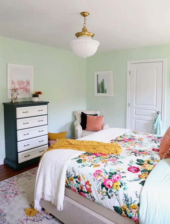 a mint colored bedroom with a bed and floral bedding, a black and white dresser, a neutral chair and a chandelier