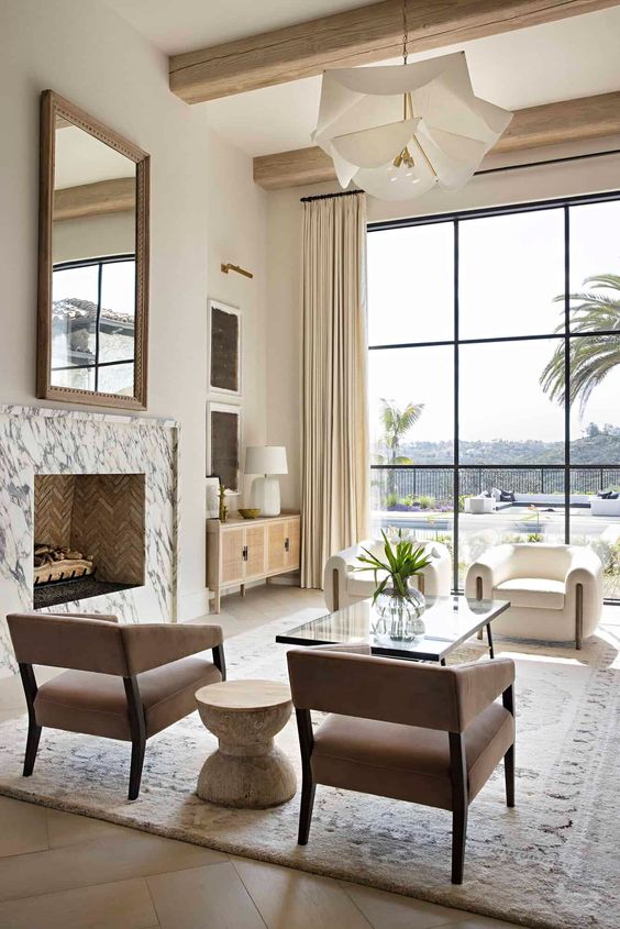 a refined Mediterranean living room done in neutral shades, with a marble clad fireplace, beige and creamy chairs, a glass coffee table and a glazed wall