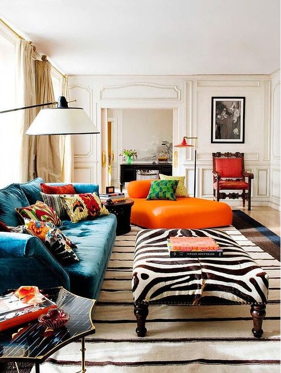 a bright maximalist living room with neutral paneled walls, a teal sofa and an orange lounger, a zebra ottoman, colorful pillows and rugs