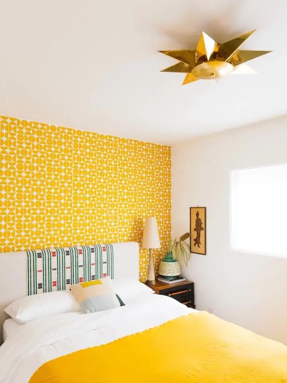 a bright bedroom with a yellow printed wallpaper wall, a neutral bed with a statement headboard and a yellow blanket, a pretty artwork is chic
