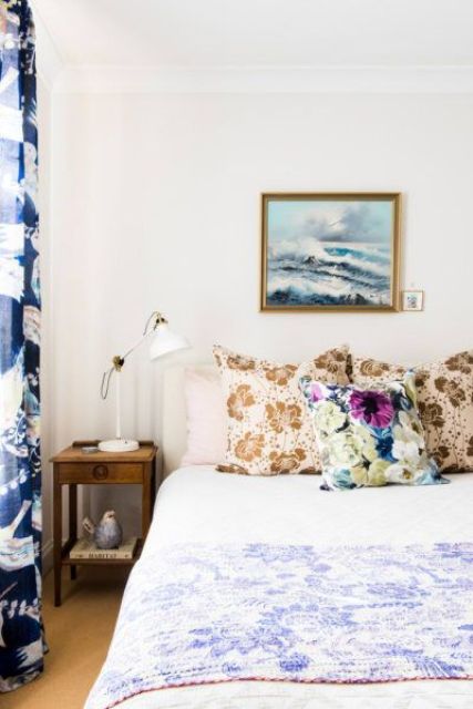 a stylish bedroom with printed pillows and bedspreads, bold printed curtains, a nightstand and a white lamp, an ocean artwork