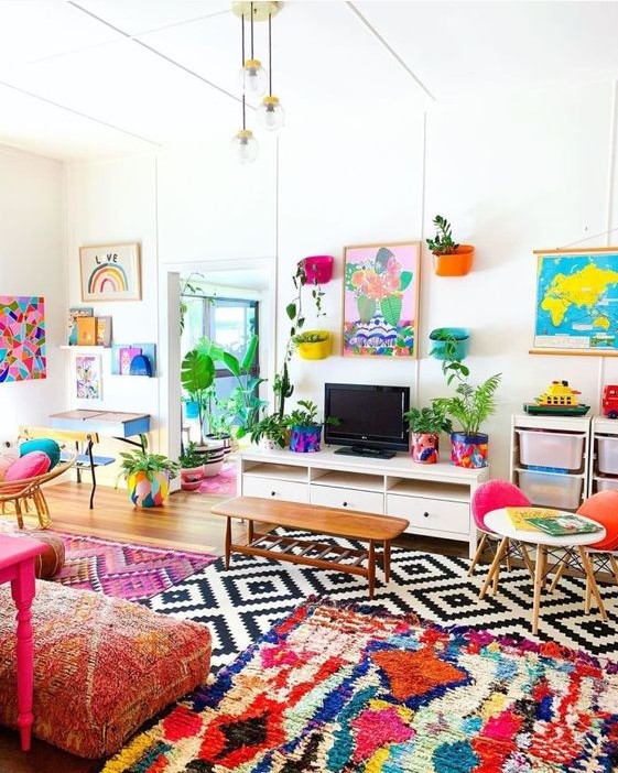 a colorful maximalist living room with bold layered rugs, pink printed cushions on the floor, some colorful chairs and artworks