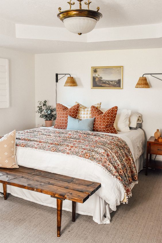 a welcoming bedroom in neutrals with a bed with floral and other printed bedding, a stained bench, a chandelier, stained nightstands