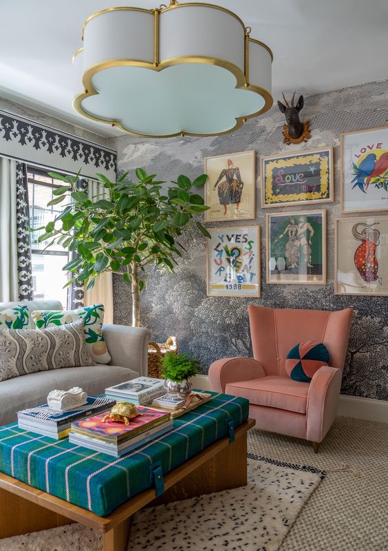 a bold maximalist living room with printed wallpaper, a grey sofa, a pink chair, an ottoman and a bright gallery wall