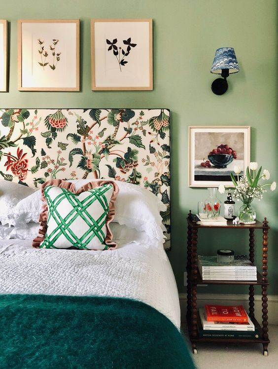 a cool and chic bedroom with green walls, a bed with a floral headboard, neutral bedding and a green bedspread, a nightstand and white blooms