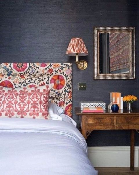 a moody bedroom with black grasscloth wallpaper, a bold floral headboard and printed pillows, a printed sconce and a stained nightstand