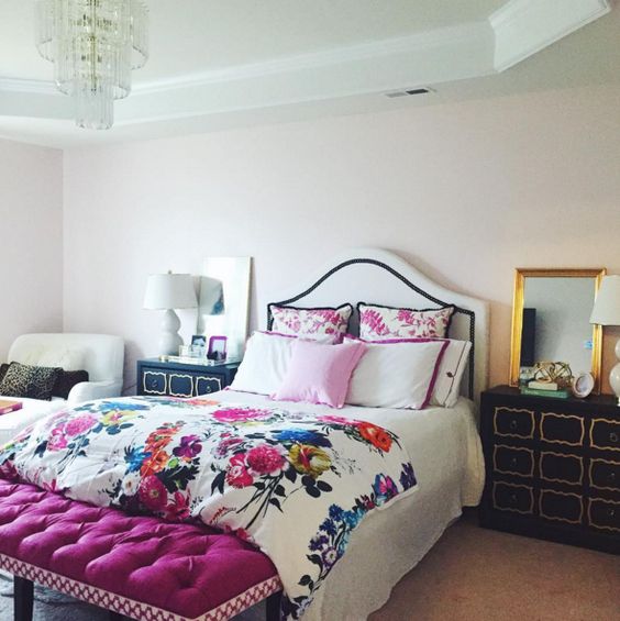 a glam bedroom with an upholstered bed and floral bedding, a fuchsia upholstered bench, black dressers and a chair