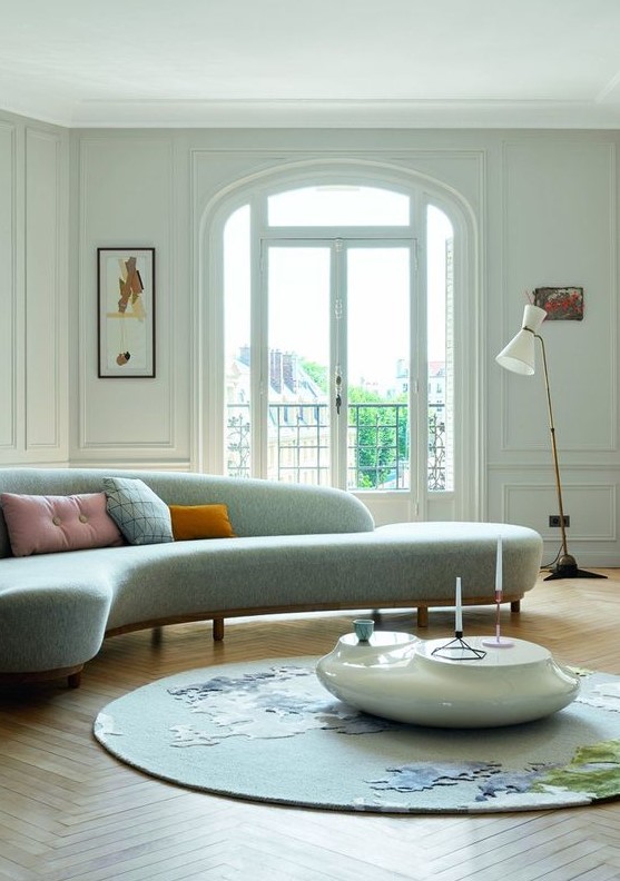 an ethereal living room with a curved blue sofa, an abstract rug and a white pebble coffee table
