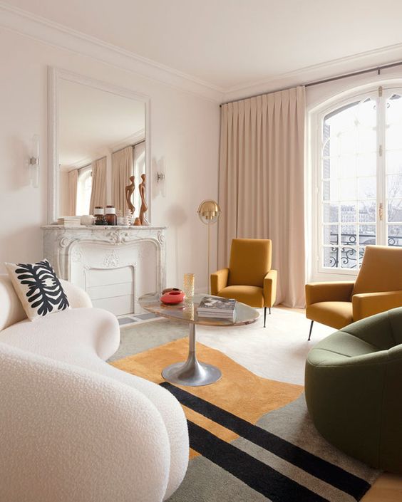 a chic living room with a faux fireplace, a creamy curved sofa, mustard and a green chairs, a catchy coffee table and a large mirror
