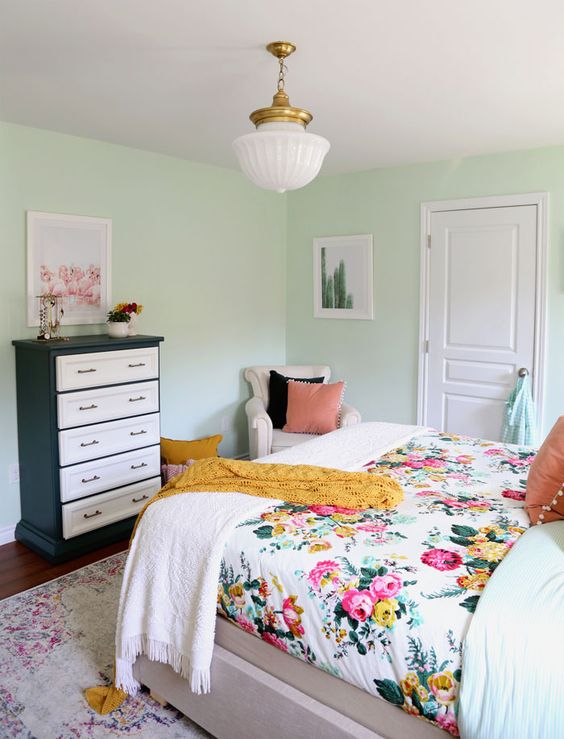 a mint-colored bedroom with a bed and floral bedding, a black and white dresser, a neutral chair and a chandelier