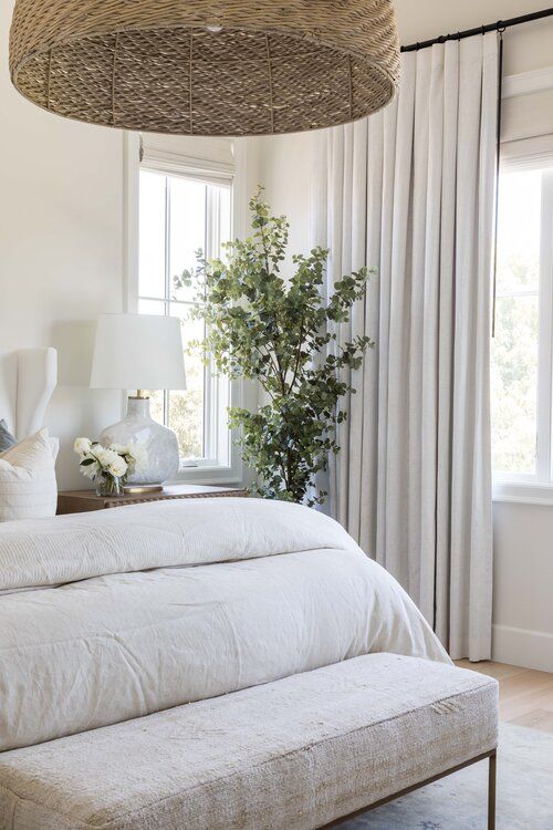 a neutral bedroom with a potted plant, neutral bedding and an upholstered bench, a woven pendant lamp