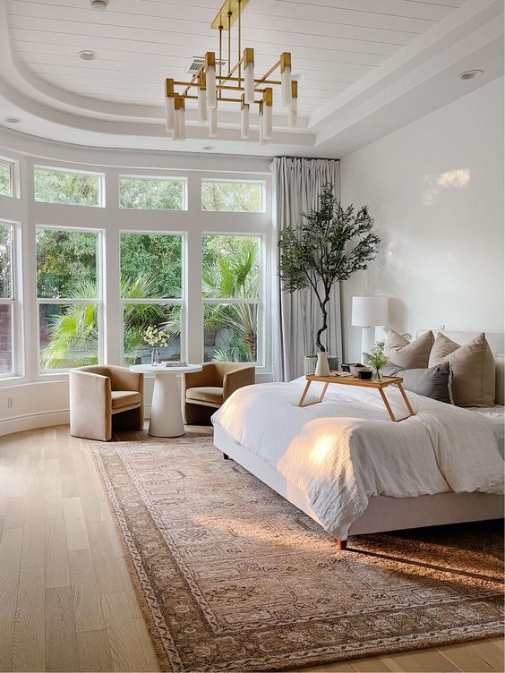 a chic neutral bedroom with an ipholstered bed, neutral bedding, a printed rug, a sitting zone and a potted plabt