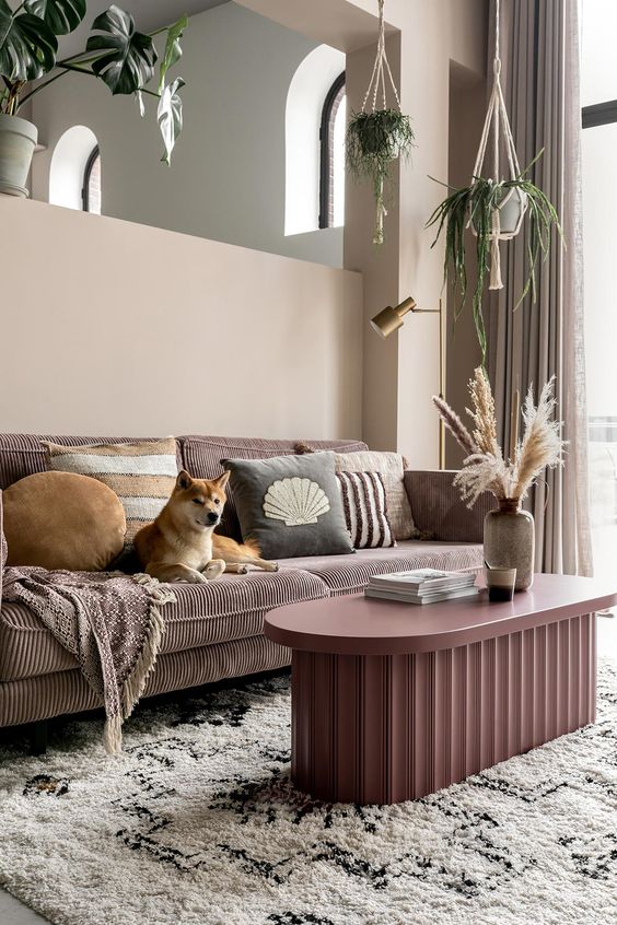a boho living room with tan walls, a mauve sofa, a pink fluted table, printed pillows and potted greenery