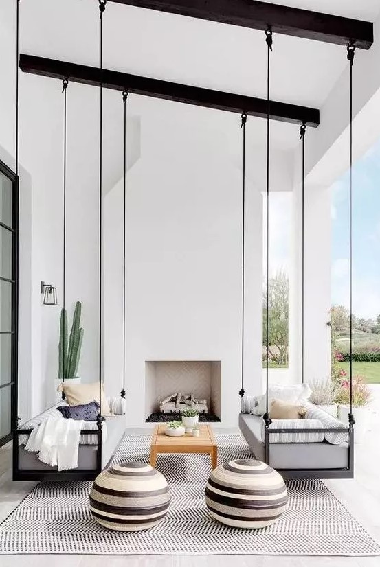 a catchy monochromatic outdoor living room with a built-in fireplace, suspended daybeds, round ottomans and a large printed rug