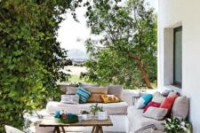 76 a lively Mediterranean outdoor space with a white built-in sofa with neutral upholstery and bright pillows, a wooden trestle table and wicker chairs, greenery weaving around the pillars