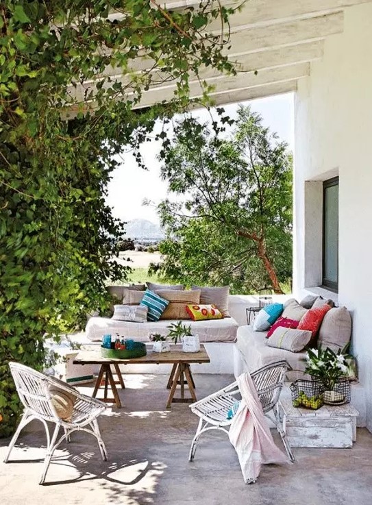 a lively Mediterranean outdoor space with a white built-in sofa with neutral upholstery and bright pillows, a wooden trestle table and wicker chairs, greenery weaving around the pillars