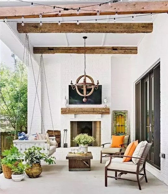 a lovely modern farmhouse outdoor living room with a fireplace, a suspended daybed, modern chairs, potted plants and blooms and a wooden chandelier